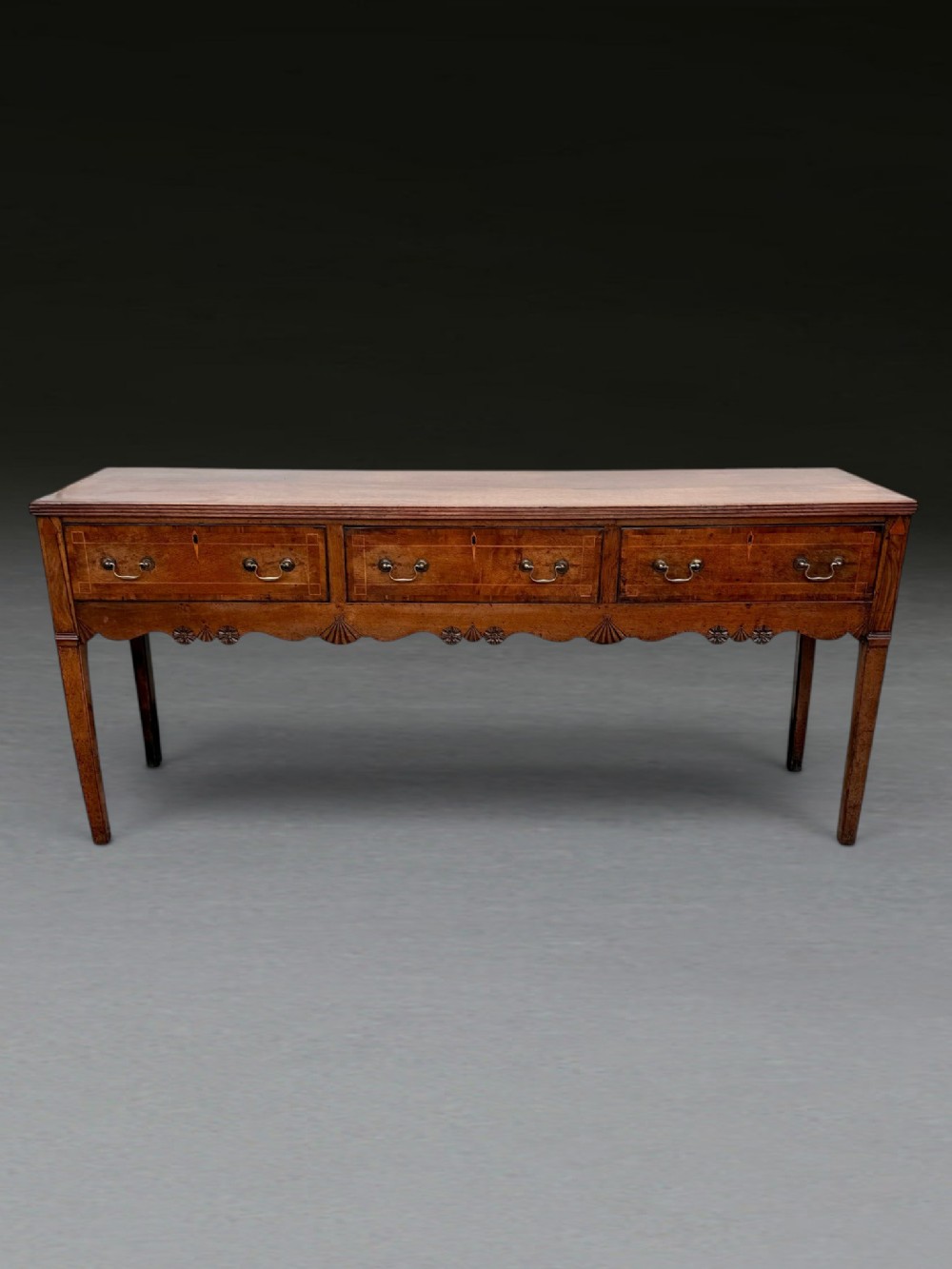 A George III oak dresser base featuring three inline drawers with brass handles, crossbanded inlay, a carved scalloped apron, and straight tapered legs.
