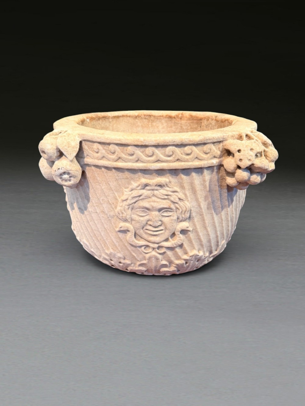A carved light buff marble urn featuring a relief face motif, fruit clusters, and diagonal fluted exterior details.