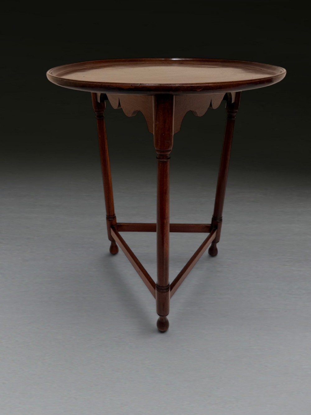 A George III mahogany cricket table with a circular top, scalloped apron, three turned legs, and a triangular stretcher base.