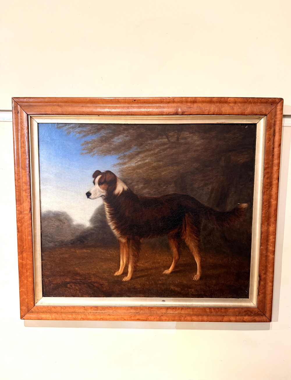 A 19th-century primitive oil painting on canvas depicting a standing brown and white dog set against a landscape backdrop, housed within a wide wood frame.