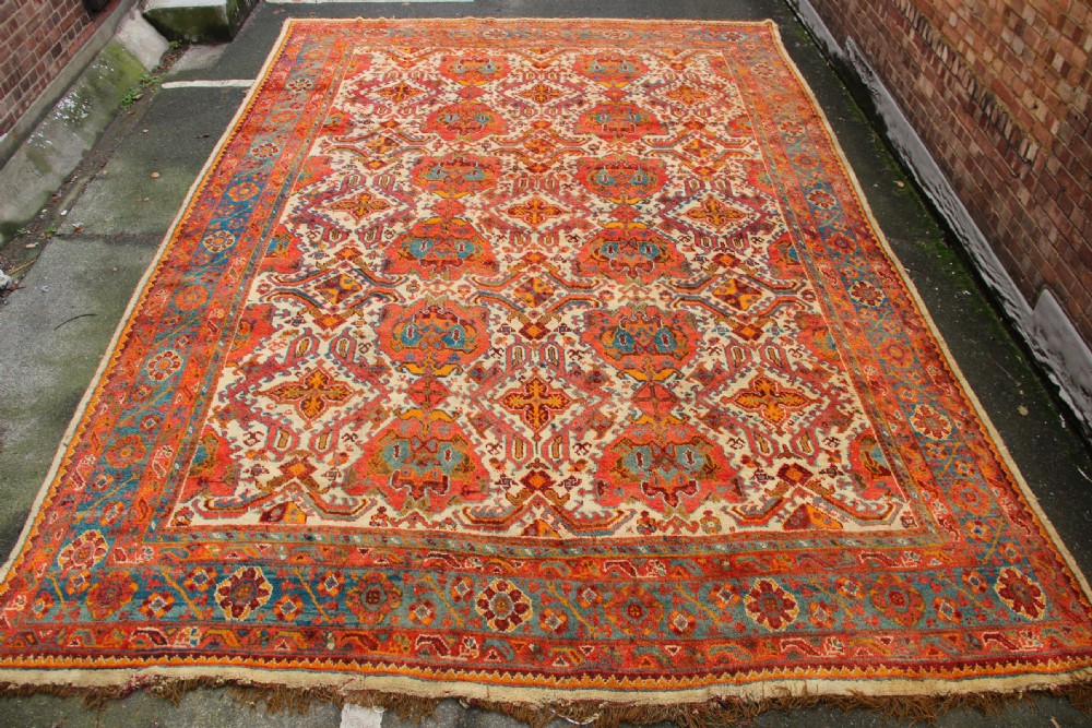 A large hand-knotted wool carpet featuring an ivory field decorated with geometric orange, terracotta, and turquoise medallion motifs within a wide multi-layered border.