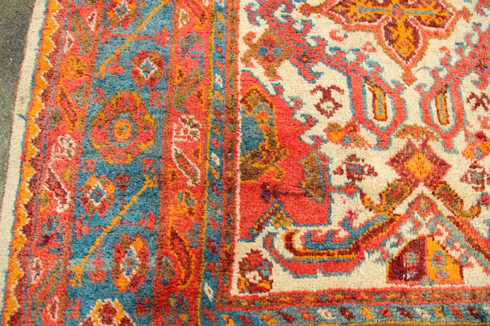 A large hand-knotted wool carpet featuring an ivory field decorated with geometric orange, terracotta, and turquoise medallion motifs within a wide multi-layered border. View V