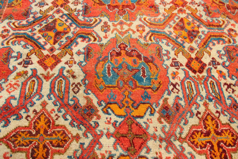 A large hand-knotted wool carpet featuring an ivory field decorated with geometric orange, terracotta, and turquoise medallion motifs within a wide multi-layered border. View VI