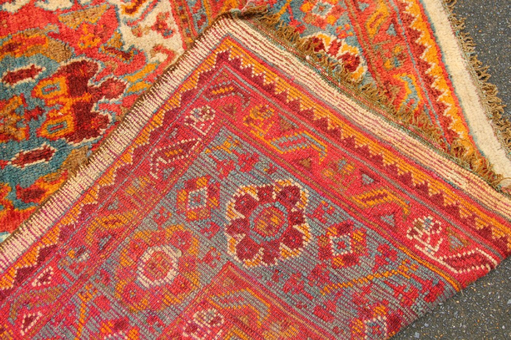 A large hand-knotted wool carpet featuring an ivory field decorated with geometric orange, terracotta, and turquoise medallion motifs within a wide multi-layered border. View IX