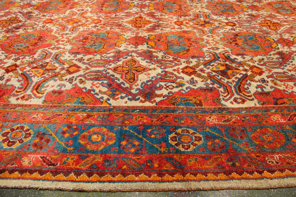 A large hand-knotted wool carpet featuring an ivory field decorated with geometric orange, terracotta, and turquoise medallion motifs within a wide multi-layered border. View XI