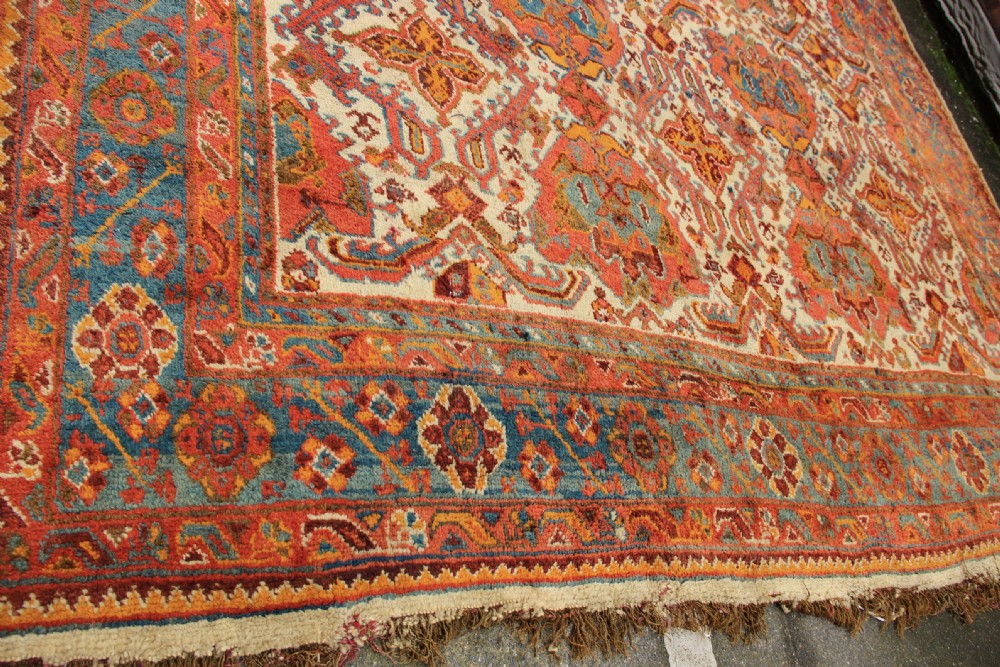 A large hand-knotted wool carpet featuring an ivory field decorated with geometric orange, terracotta, and turquoise medallion motifs within a wide multi-layered border. View XIII