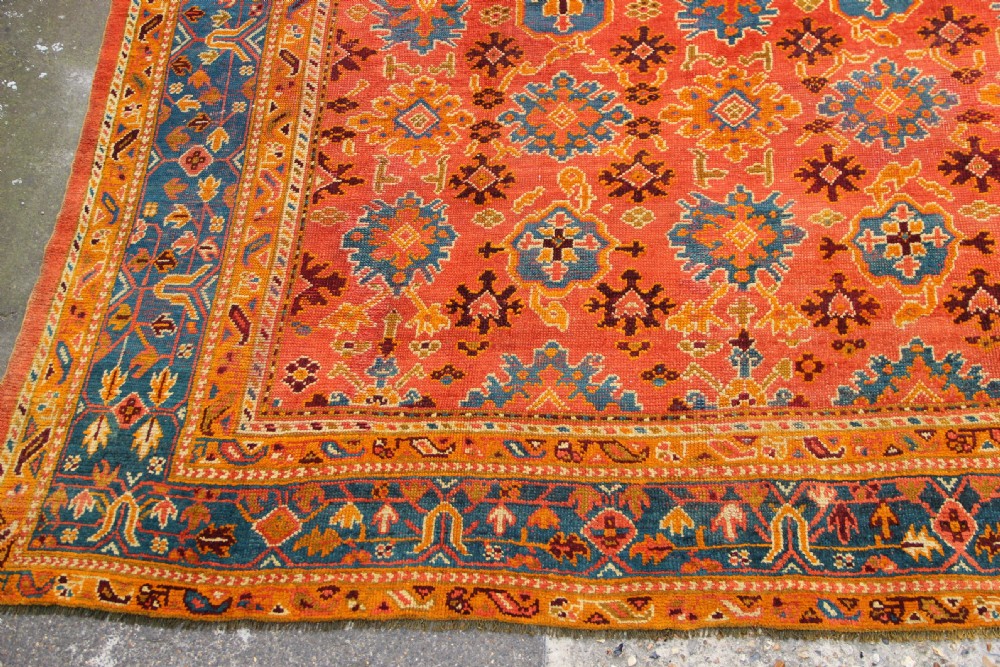 A large square antique Oushak carpet featuring a terracotta orange field with repeated blue and yellow geometric floral medallions within a multi-tiered border. View II