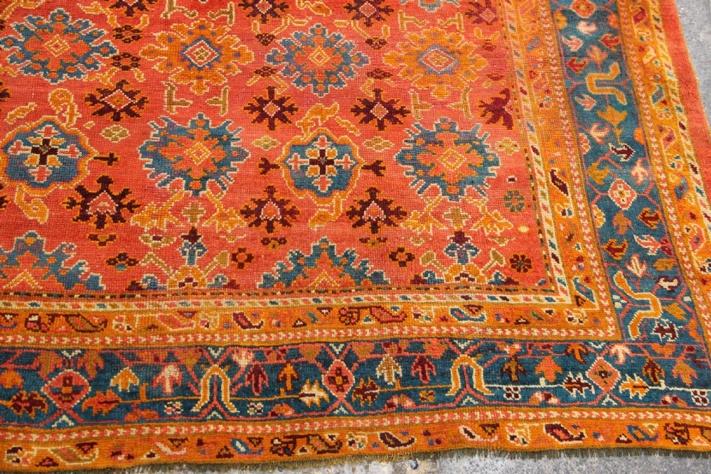 A large square antique Oushak carpet featuring a terracotta orange field with repeated blue and yellow geometric floral medallions within a multi-tiered border. View III