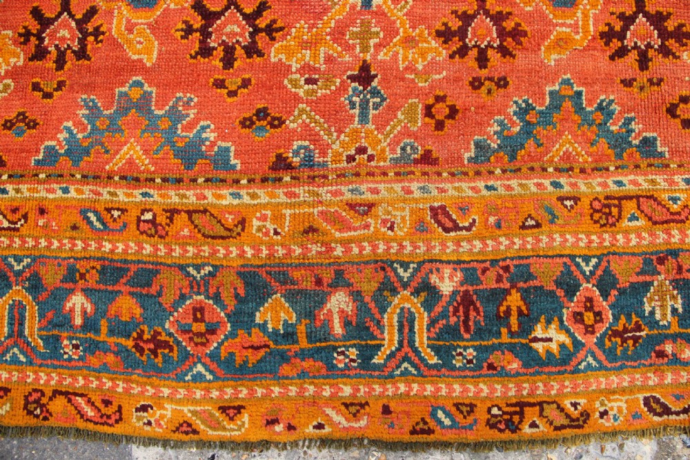 A large square antique Oushak carpet featuring a terracotta orange field with repeated blue and yellow geometric floral medallions within a multi-tiered border. View V