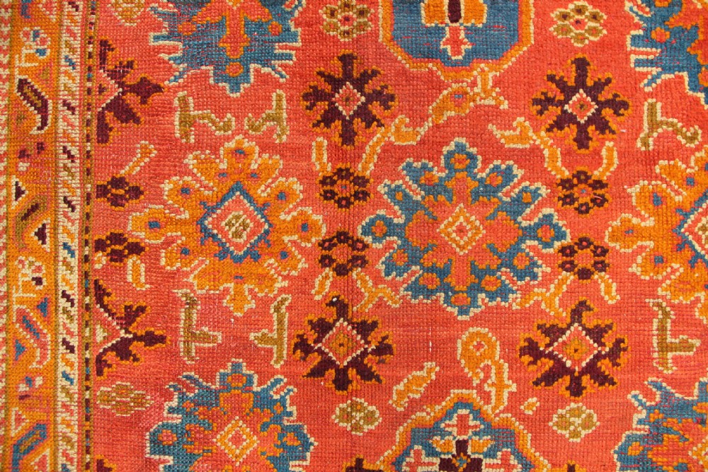 A large square antique Oushak carpet featuring a terracotta orange field with repeated blue and yellow geometric floral medallions within a multi-tiered border. View VI
