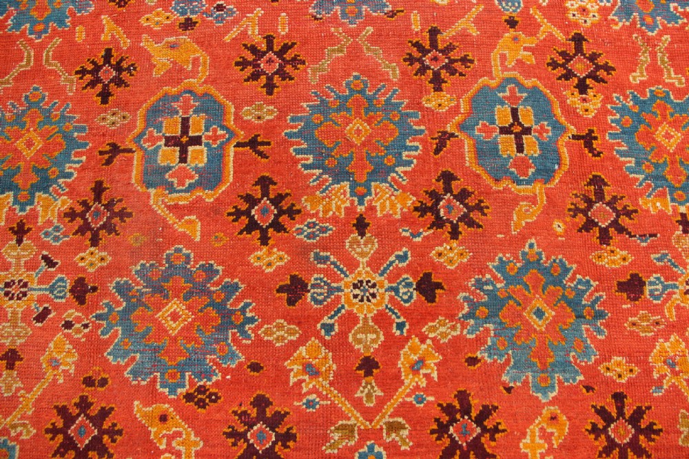 A large square antique Oushak carpet featuring a terracotta orange field with repeated blue and yellow geometric floral medallions within a multi-tiered border. View VII