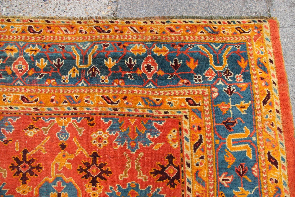 A large square antique Oushak carpet featuring a terracotta orange field with repeated blue and yellow geometric floral medallions within a multi-tiered border. View VIII