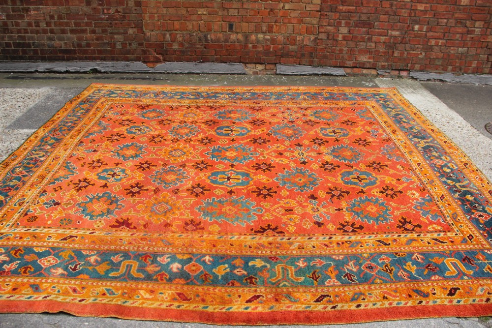 A large square antique Oushak carpet featuring a terracotta orange field with repeated blue and yellow geometric floral medallions within a multi-tiered border. View IX