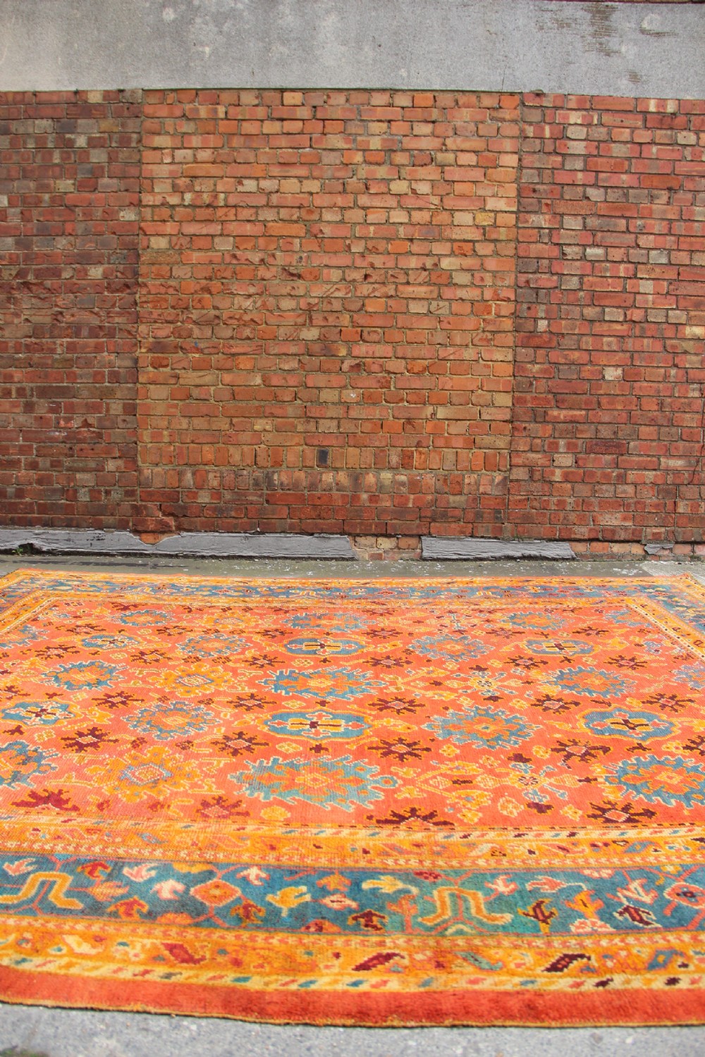 A large square antique Oushak carpet featuring a terracotta orange field with repeated blue and yellow geometric floral medallions within a multi-tiered border. View X