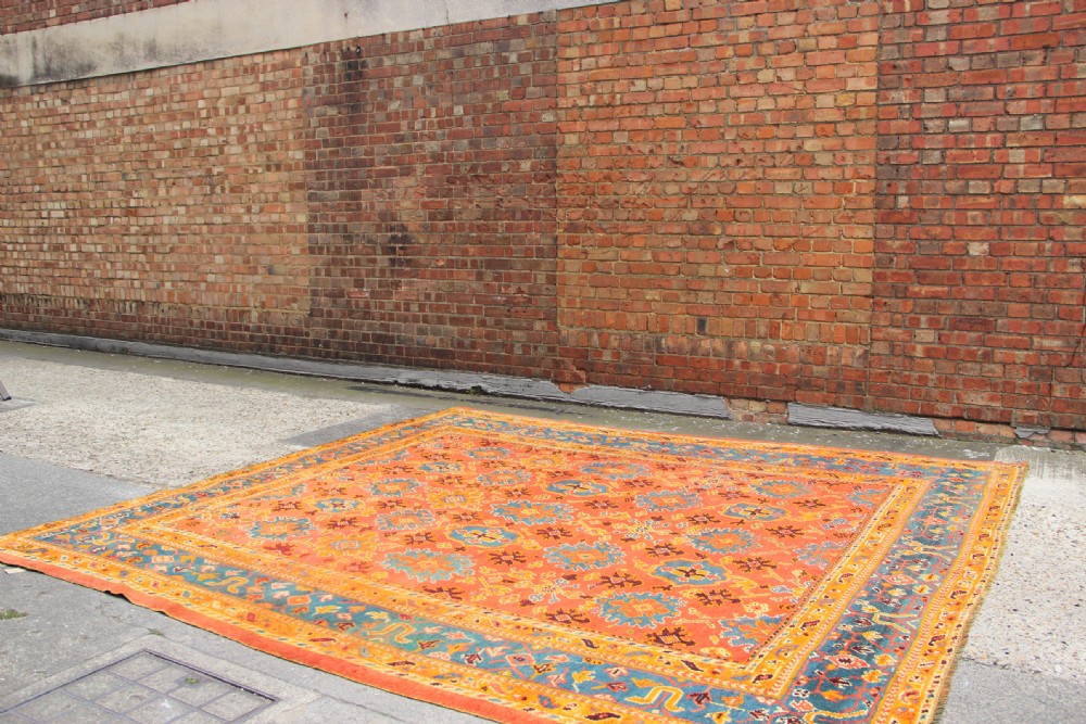 A large square antique Oushak carpet featuring a terracotta orange field with repeated blue and yellow geometric floral medallions within a multi-tiered border. View XI