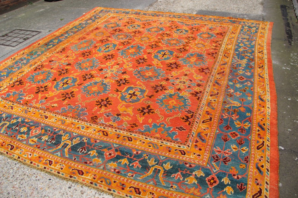 A large square antique Oushak carpet featuring a terracotta orange field with repeated blue and yellow geometric floral medallions within a multi-tiered border. View XII