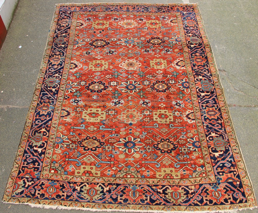 An antique Heriz wool carpet featuring a terracotta red field filled with geometric floral motifs framed by a navy blue border.
