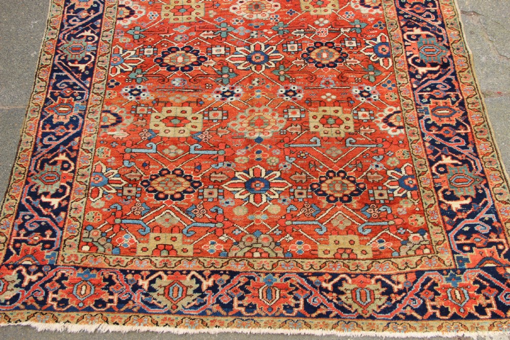 An antique Heriz wool carpet featuring a terracotta red field filled with geometric floral motifs framed by a navy blue border. View II