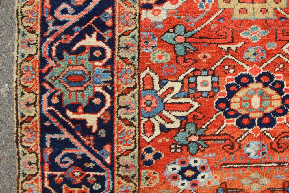 An antique Heriz wool carpet featuring a terracotta red field filled with geometric floral motifs framed by a navy blue border. View V