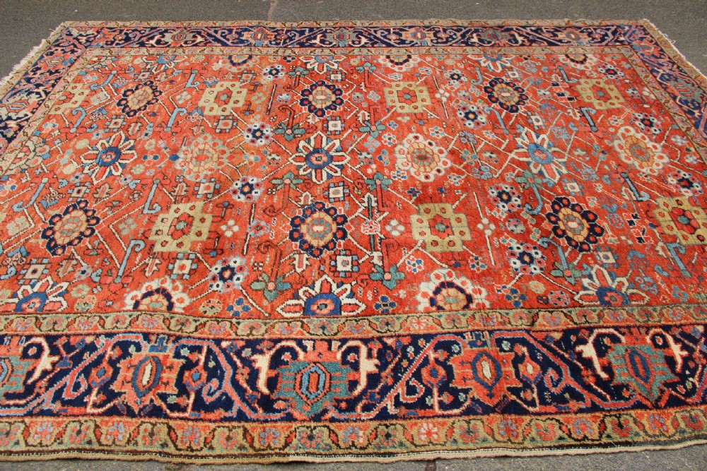 An antique Heriz wool carpet featuring a terracotta red field filled with geometric floral motifs framed by a navy blue border. View VII