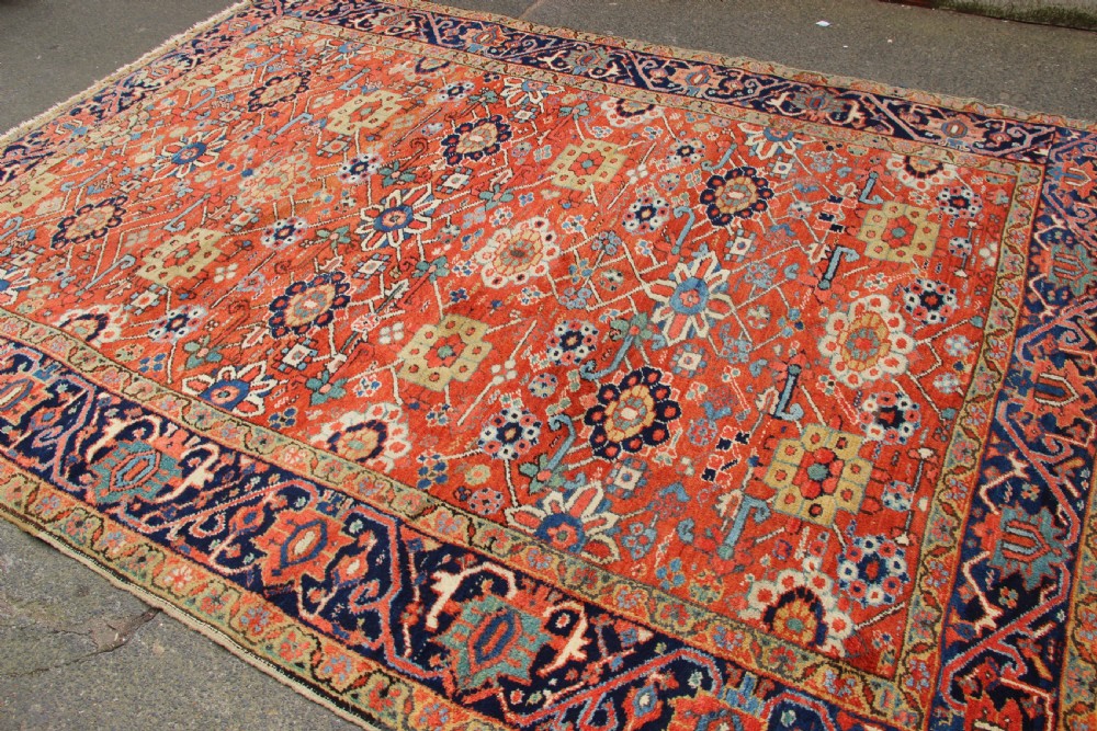 An antique Heriz wool carpet featuring a terracotta red field filled with geometric floral motifs framed by a navy blue border. View VIII