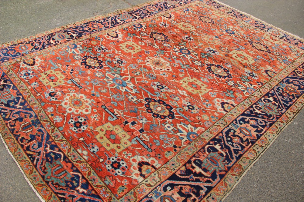 An antique Heriz wool carpet featuring a terracotta red field filled with geometric floral motifs framed by a navy blue border. View IX