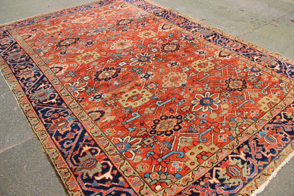 An antique Heriz wool carpet featuring a terracotta red field filled with geometric floral motifs framed by a navy blue border. View X