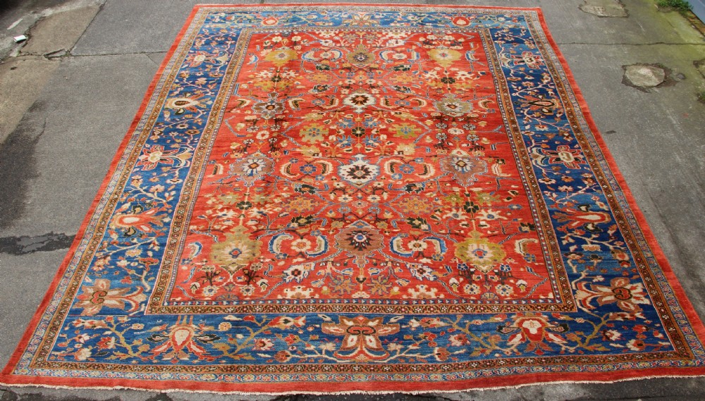 A large hand-knotted antique Ziegler carpet featuring a terracotta red field with floral palmettes framed by a wide indigo blue border.