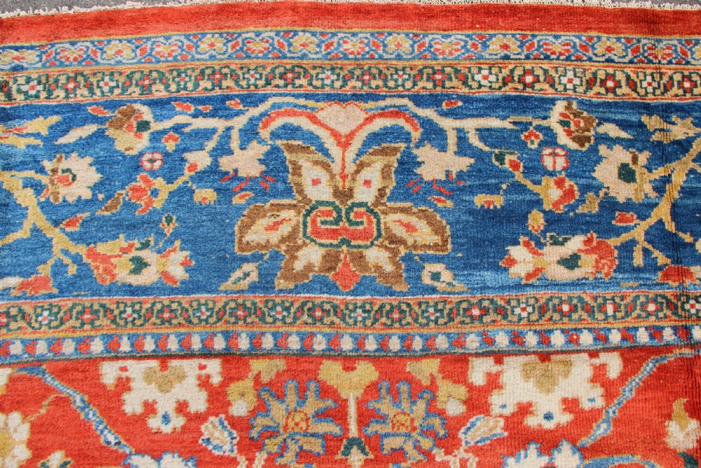 A large hand-knotted antique Ziegler carpet featuring a terracotta red field with floral palmettes framed by a wide indigo blue border. View VIII