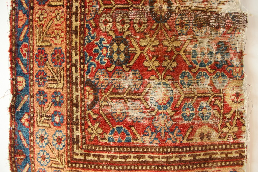 An antique hand-knotted textile fragment displaying geometric patterns and floral motifs in shades of terracotta red, indigo blue, and cream. View III