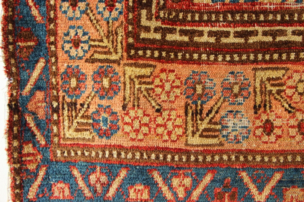 An antique hand-knotted textile fragment displaying geometric patterns and floral motifs in shades of terracotta red, indigo blue, and cream. View IV