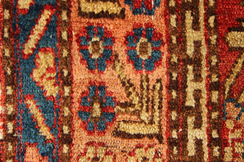 An antique hand-knotted textile fragment displaying geometric patterns and floral motifs in shades of terracotta red, indigo blue, and cream. View V