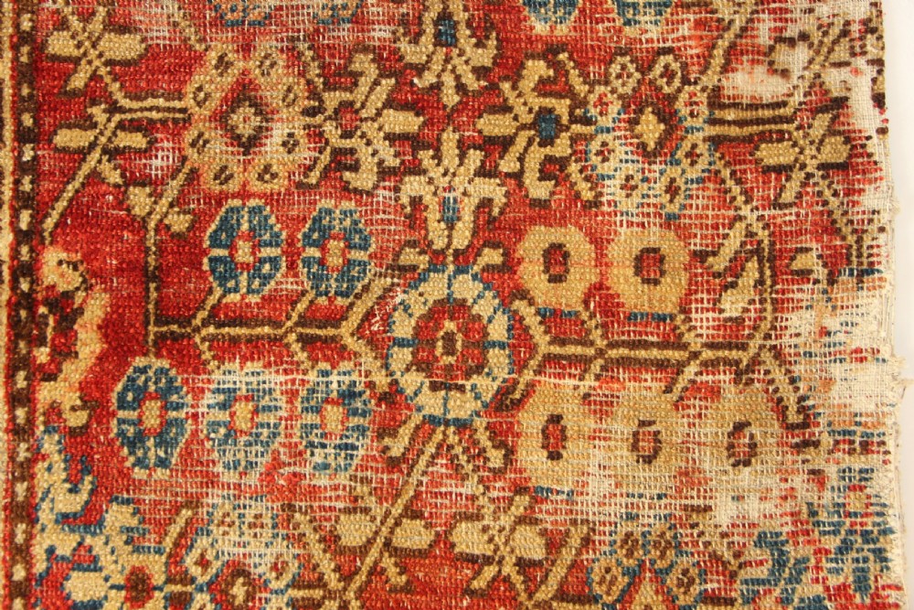 An antique hand-knotted textile fragment displaying geometric patterns and floral motifs in shades of terracotta red, indigo blue, and cream. View VI