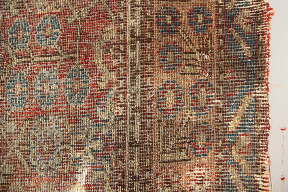 An antique hand-knotted textile fragment displaying geometric patterns and floral motifs in shades of terracotta red, indigo blue, and cream. View VII