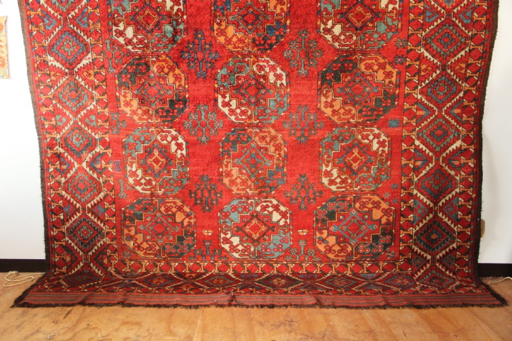 A large hand-knotted wool carpet featuring repeating octagonal gul medallions and geometric borders in terracotta red, navy blue, and cream tones. View III