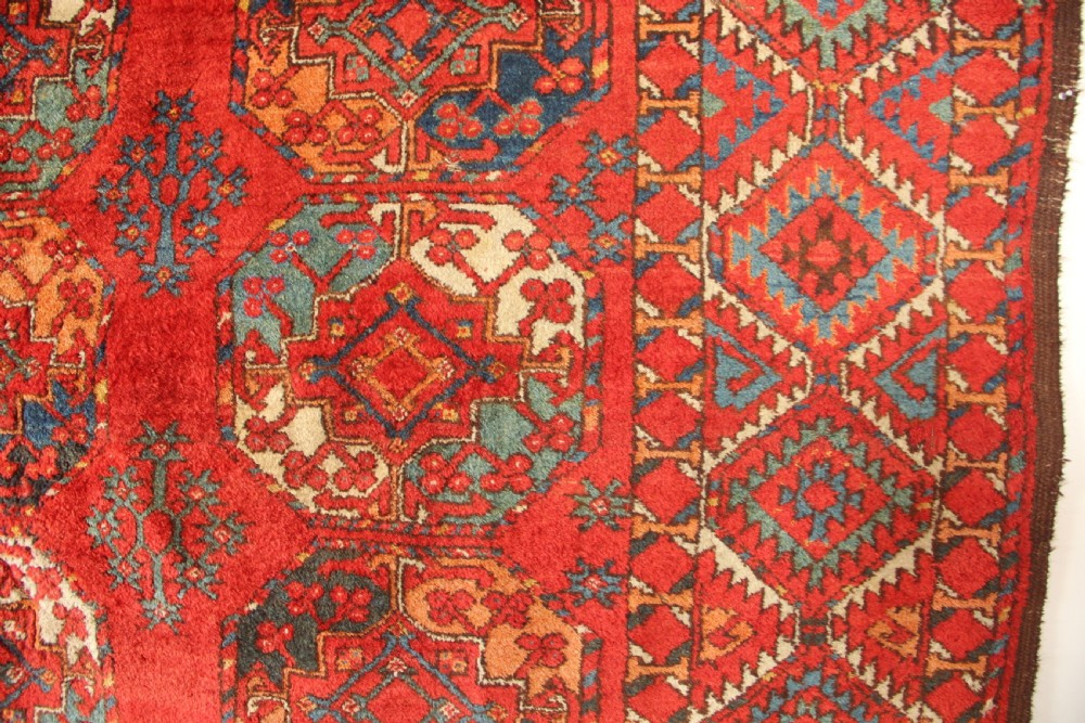 A large hand-knotted wool carpet featuring repeating octagonal gul medallions and geometric borders in terracotta red, navy blue, and cream tones. View IV
