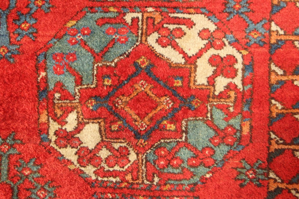 A large hand-knotted wool carpet featuring repeating octagonal gul medallions and geometric borders in terracotta red, navy blue, and cream tones. View V