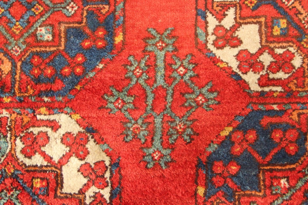 A large hand-knotted wool carpet featuring repeating octagonal gul medallions and geometric borders in terracotta red, navy blue, and cream tones. View VI