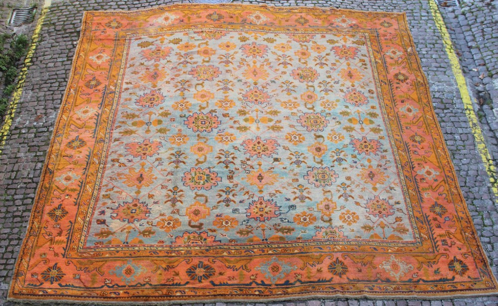 A large square antique Oushak rug with a light blue central field decorated with floral medallions and a wide terracotta and orange floral border.