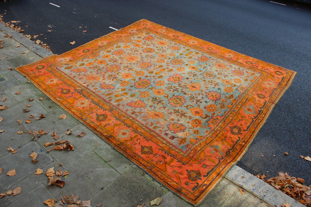 A large square antique Oushak rug with a light blue central field decorated with floral medallions and a wide terracotta and orange floral border. View II