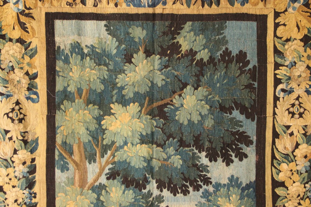An 18th-century vertical verdure tapestry depicting a woodland landscape with a bird and lush trees framed by a decorative floral woven border. View V
