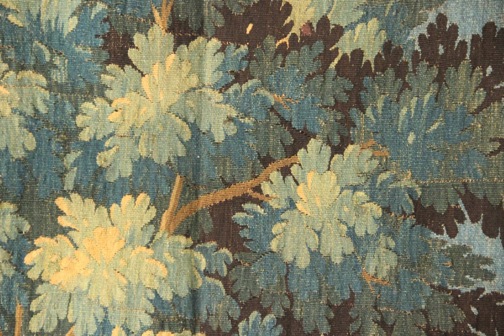 An 18th-century vertical verdure tapestry depicting a woodland landscape with a bird and lush trees framed by a decorative floral woven border. View VI