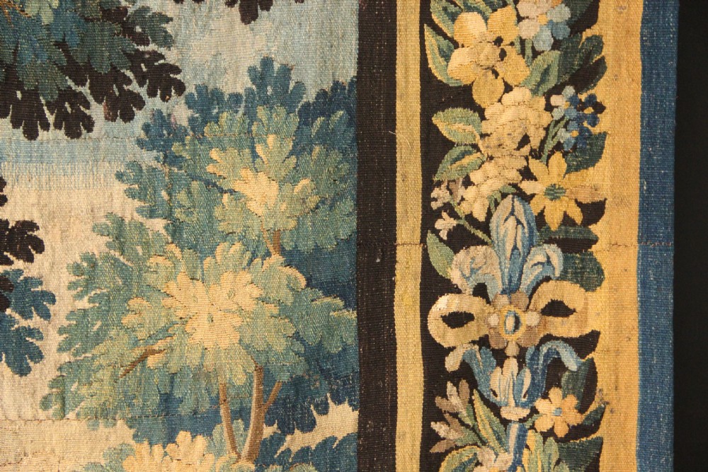 An 18th-century vertical verdure tapestry depicting a woodland landscape with a bird and lush trees framed by a decorative floral woven border. View VII