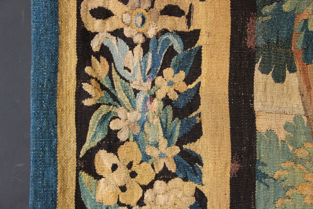 An 18th-century vertical verdure tapestry depicting a woodland landscape with a bird and lush trees framed by a decorative floral woven border. View VIII