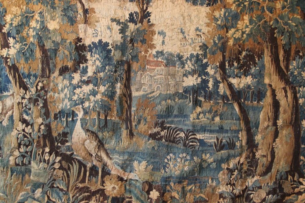 A large 18th-century hand-woven verdure tapestry depicting a dense woodland scene with foliage, animals, and distant architecture within a decorative floral border. View II