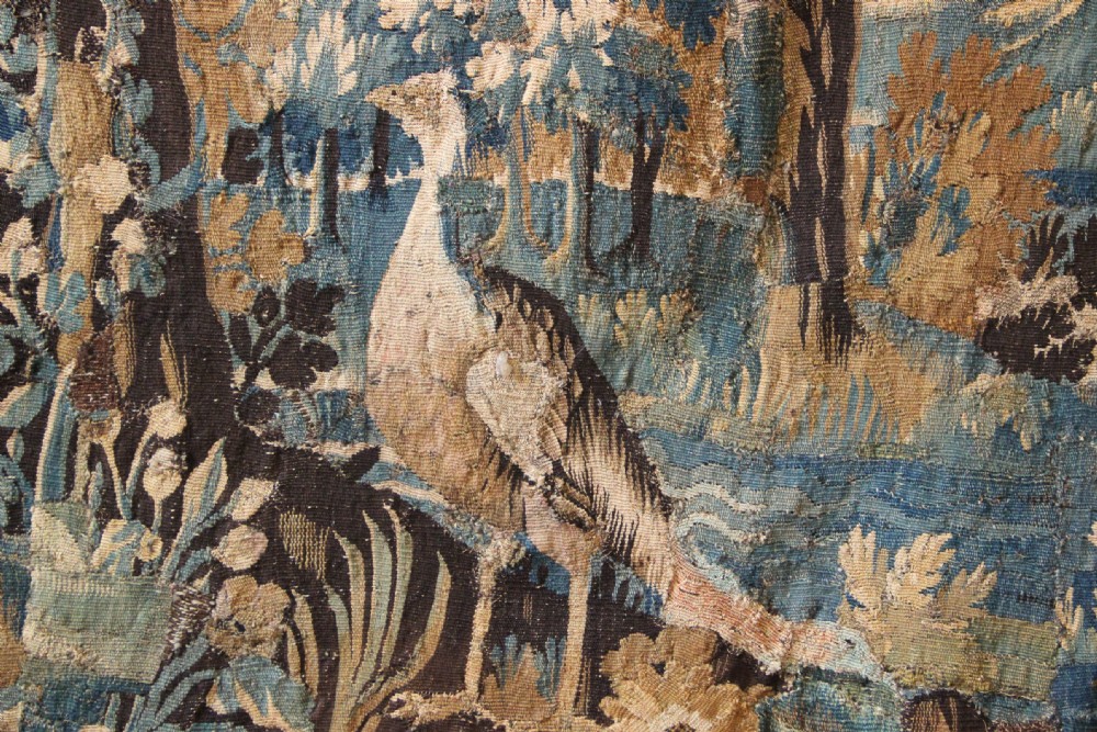 A large 18th-century hand-woven verdure tapestry depicting a dense woodland scene with foliage, animals, and distant architecture within a decorative floral border. View V