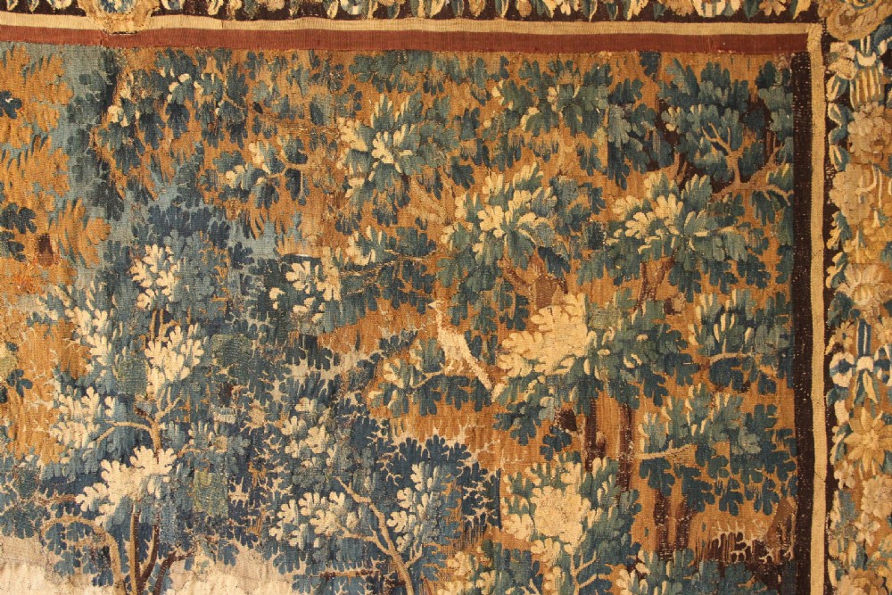A large 18th-century hand-woven verdure tapestry depicting a dense woodland scene with foliage, animals, and distant architecture within a decorative floral border. View VI