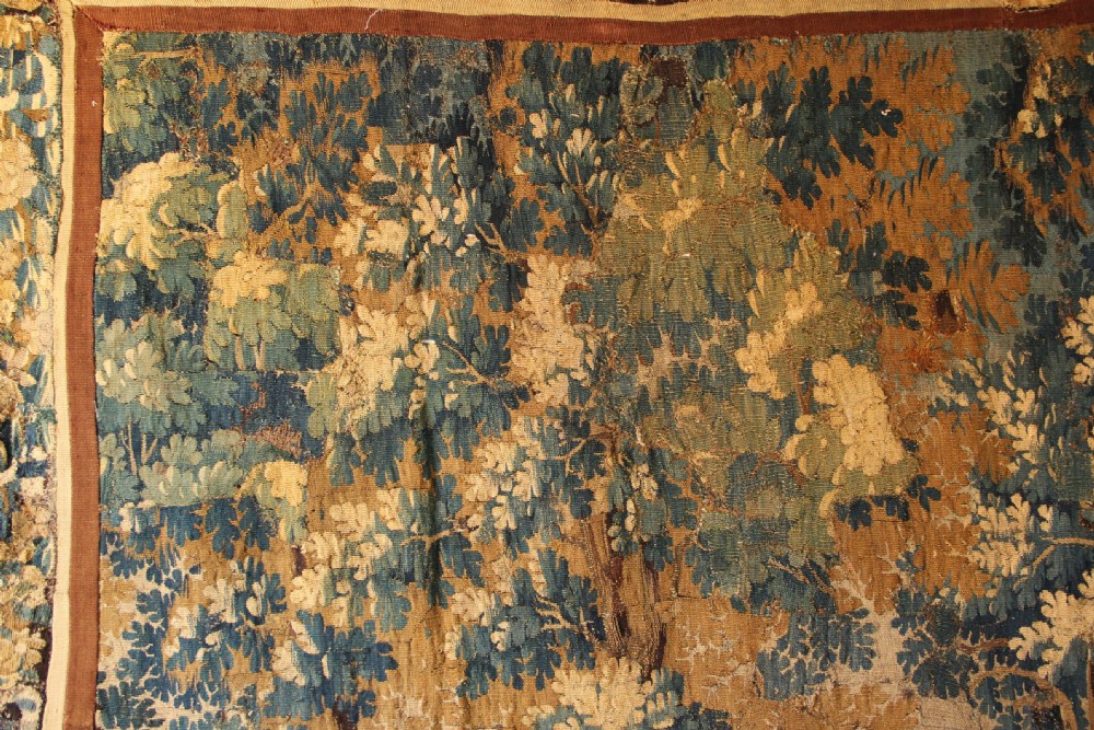 A large 18th-century hand-woven verdure tapestry depicting a dense woodland scene with foliage, animals, and distant architecture within a decorative floral border. View VII