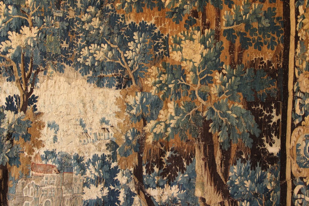 A large 18th-century hand-woven verdure tapestry depicting a dense woodland scene with foliage, animals, and distant architecture within a decorative floral border. View VIII