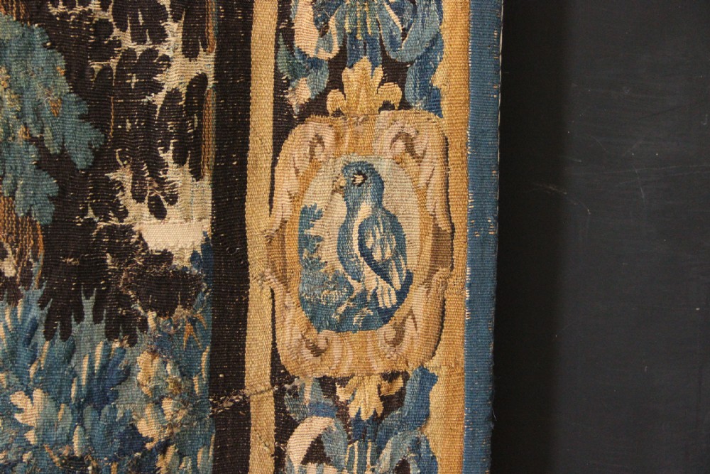 A large 18th-century hand-woven verdure tapestry depicting a dense woodland scene with foliage, animals, and distant architecture within a decorative floral border. View IX